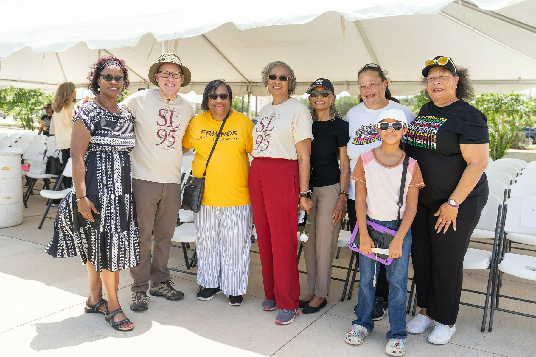 A group of eight people pose in front of a temporary event tent, several in tee-shirts with messages about Juneteenth and the Sugar Land 95.
